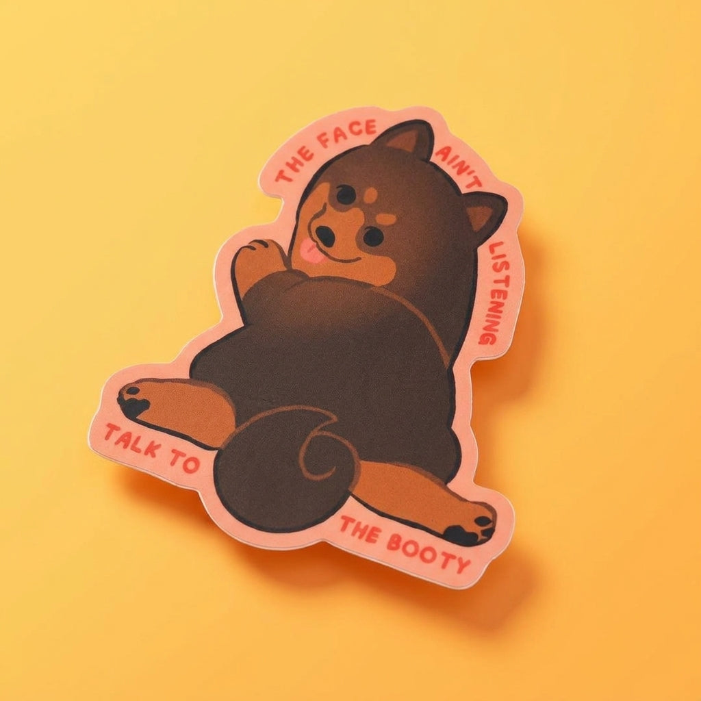 Angled Talk to the Booty Shiba die-cut waterproof vinyl sticker photographed at a three-quarter angle on a warm yellow background with a soft orange shadow.