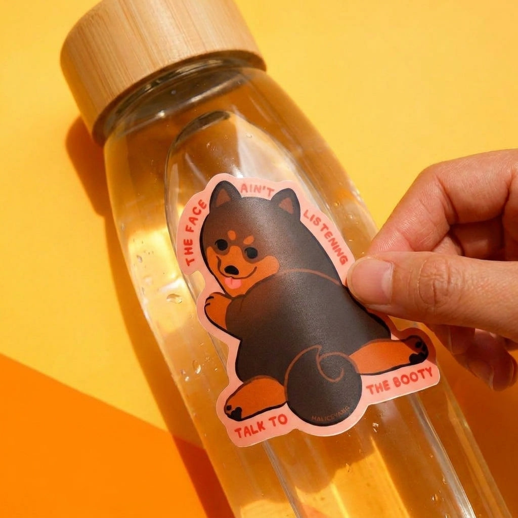 Talk to the Booty Shiba waterproof vinyl sticker being applied to a clear reusable water bottle against a warm yellow background.